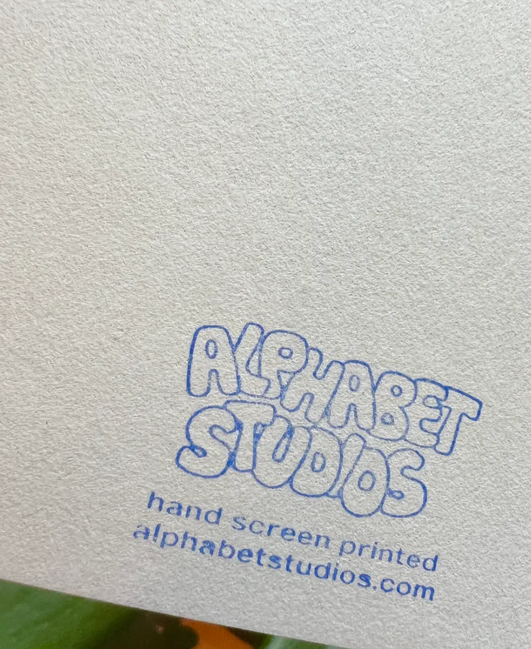 A close-up of textured off-white paper with blue hand-screen printed text reading 'ALPHABET STUDIOS' and smaller text below reading 'hand screen printed alphabetstudios.com'.
