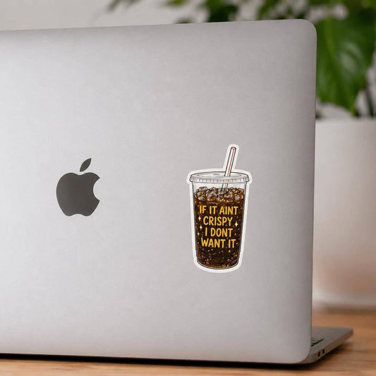 A die-cut vinyl sticker of a iced coffee cup with the text 'IF IT AIN'T CRISPY, I DON'T WANT IT' applied to the back of a silver laptop. The photo is taken on a wooden desk with a blurred background showing a plant and a candle.
