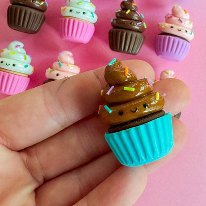 Little cupcake magnet with a teal cup and chocolate frosting being held in an open hand. Many magnets in the background.