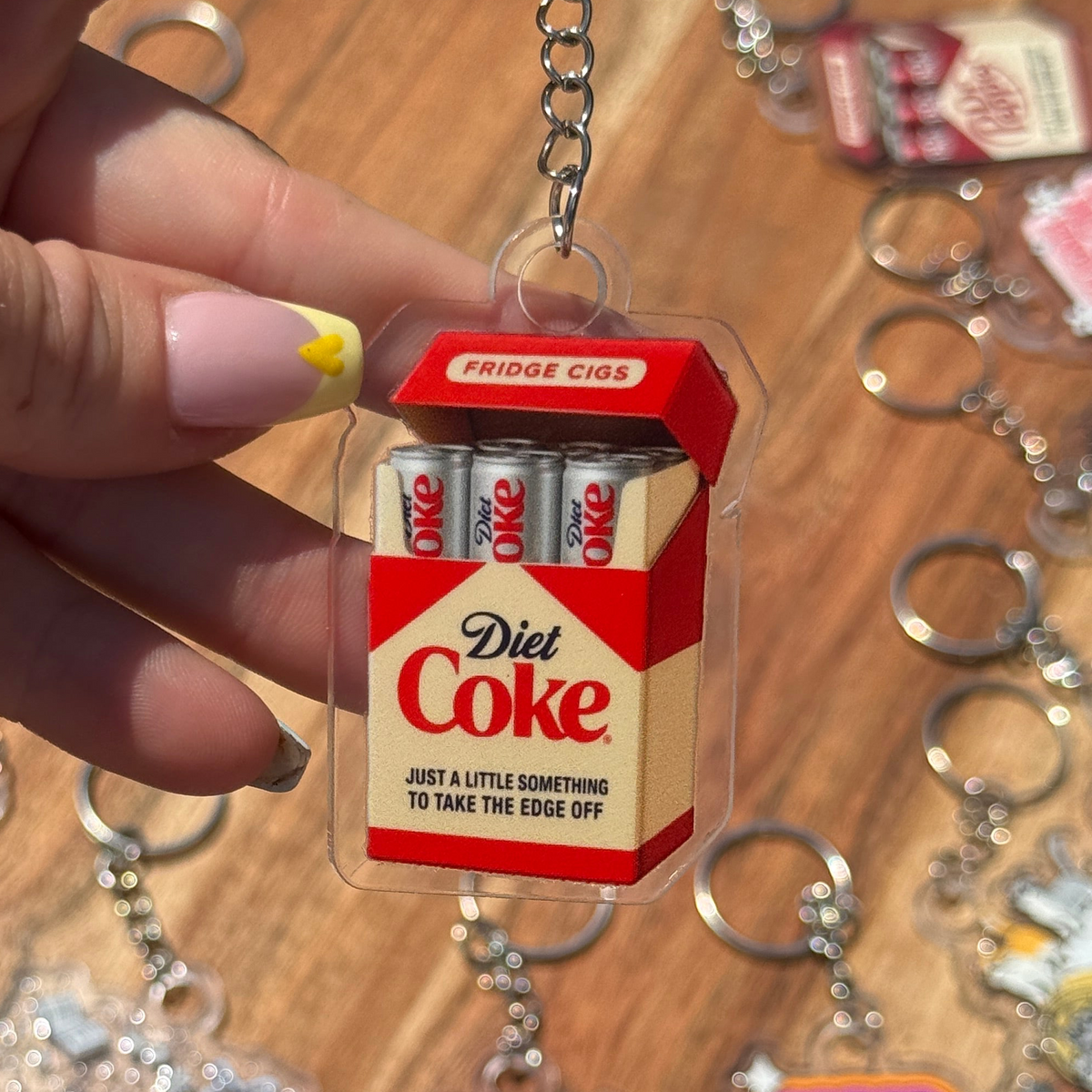 A hand holds a novelty keychain shaped like a Diet Coke Fridge Cigarettes pack, featuring clear plastic, red and beige colors, and silver metal key rings, with other keychains blurred in the background on a wooden table.