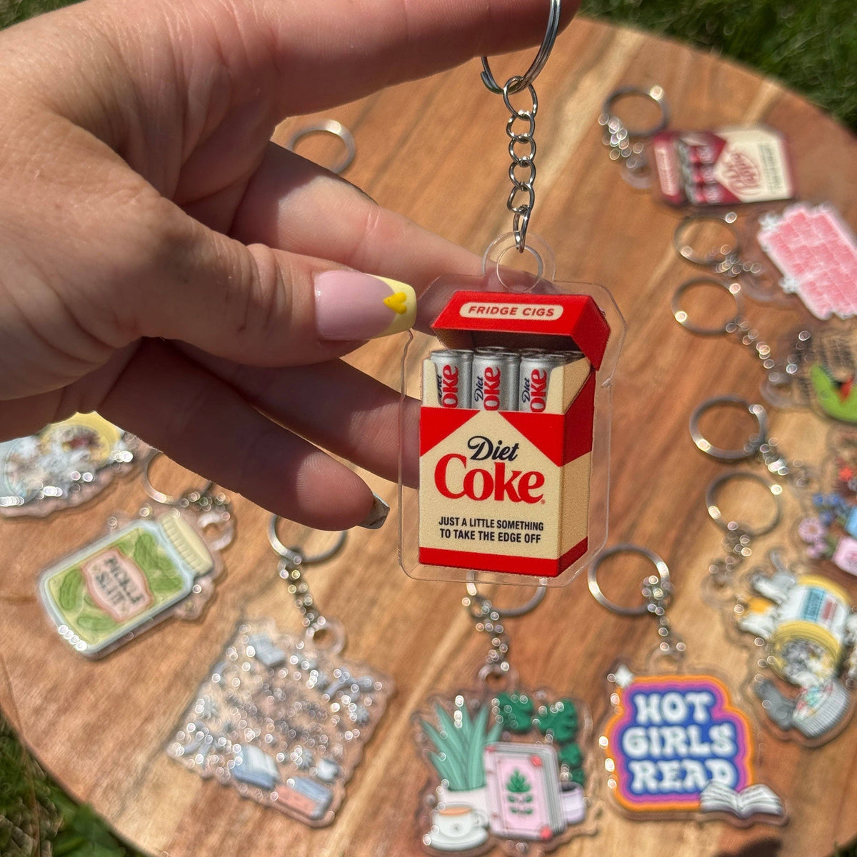 A hand holds a Diet Coke-themed keychain shaped like a miniature red and white cigarette pack with three batteries. The keychain is shown outdoors on a round wooden tray with several other colorful keychains in the background on grass.