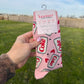 A hand holds a pair of pink patterned socks on a plastic hanger outdoors in a grassy field under a blue sky. The socks feature a retro Coke bottle graphic print with the words 'Coke' and 'Come on down to the bar.