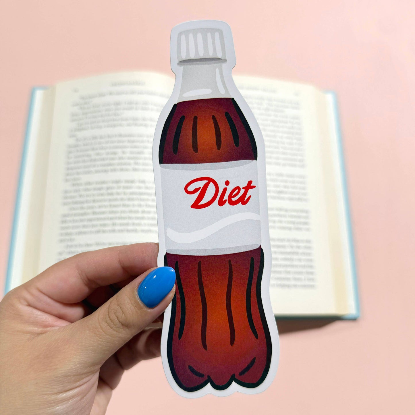 Hand holding a Diet Coke bottle-shaped bookmark with an open book in the background.