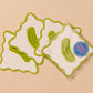 Four square white coasters with wavy green borders and green pickle illustrations, with one coaster still in clear packaging showing a blue circular label, on a warm beige background.