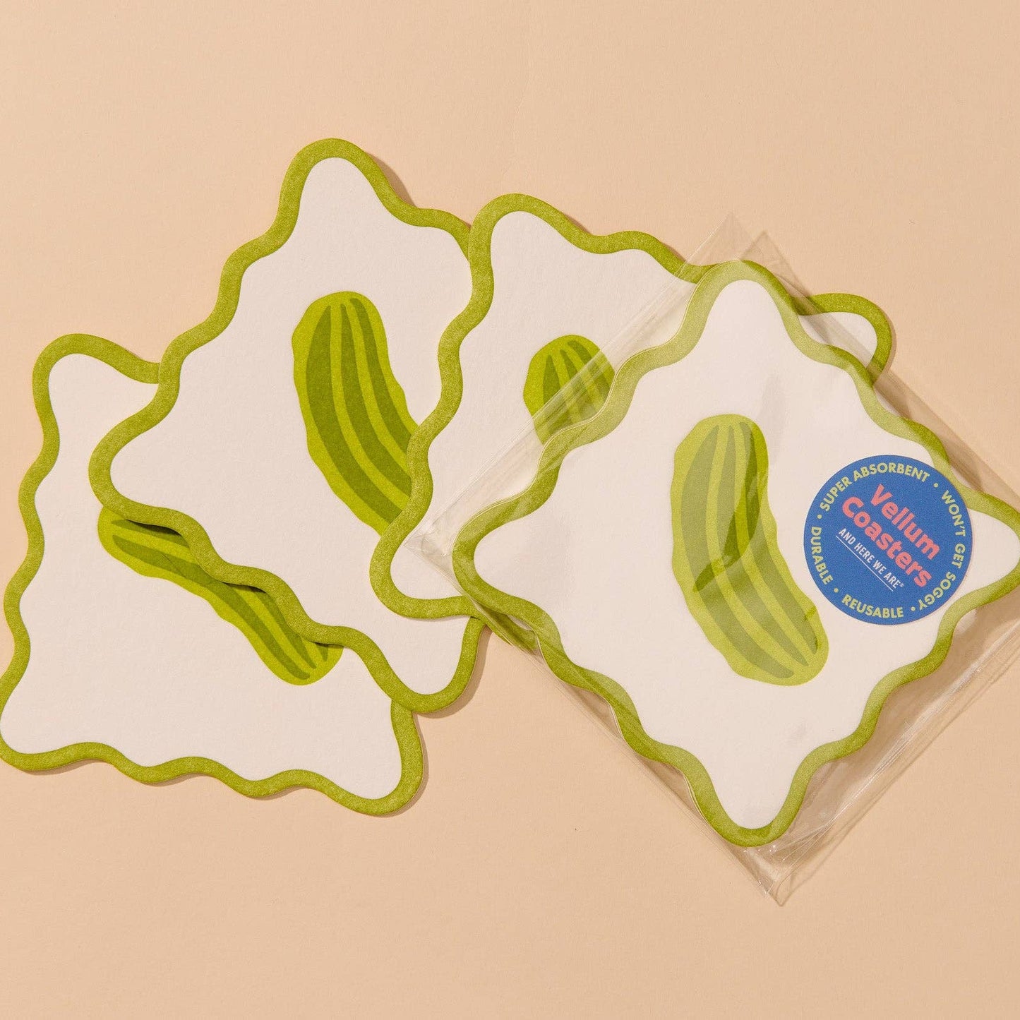Four square white coasters with wavy green borders and green pickle illustrations, with one coaster still in clear packaging showing a blue circular label, on a warm beige background.