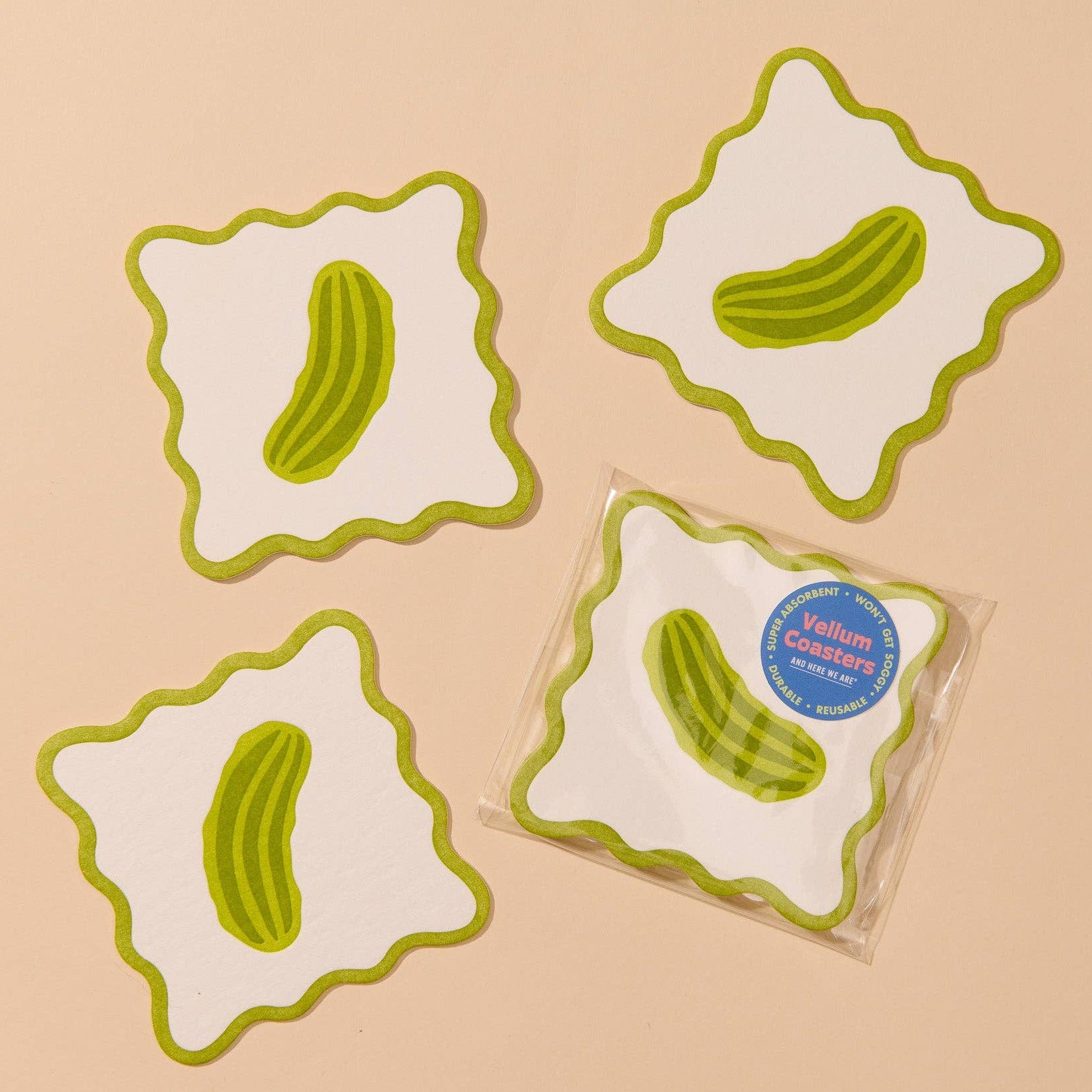 Four white square coasters with wavy green edges, each printed with a green pickle illustration, arranged on a light beige background. One coaster is shown in clear packaging with a blue circular sticker that reads 'Vellum coasters'.
