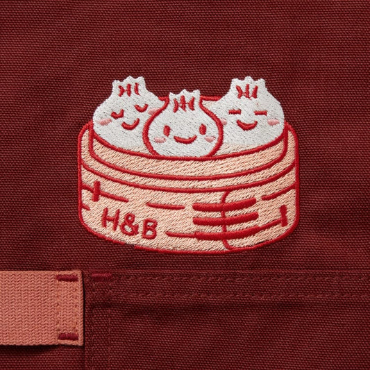 Cinnamon colored apron with mauve straps. This apron has a design that is dedicated to Dim Sum and has a steamer basket full of smiling dumplings and one dumpling being grabbed by chopsticks.