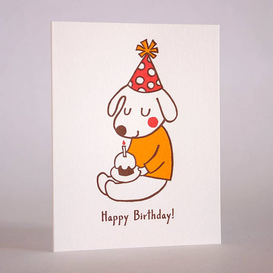 Card with a dog wearing a polkda dot party hat and a mustard colored sweater. Dog is holding a cupcake that has a candle in it. Text reads: Happy Birthday!