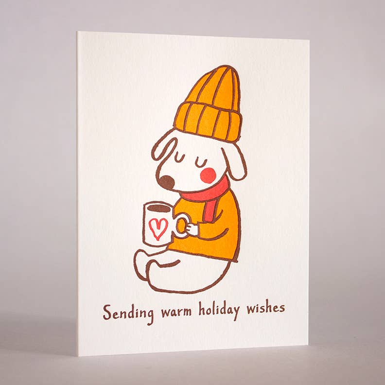 Dog wearing a mustard colored beanie and sweater with red scarf sitting with a cup of warm liquid. Text reads: Sending warm holiday wishes. Greeting card.