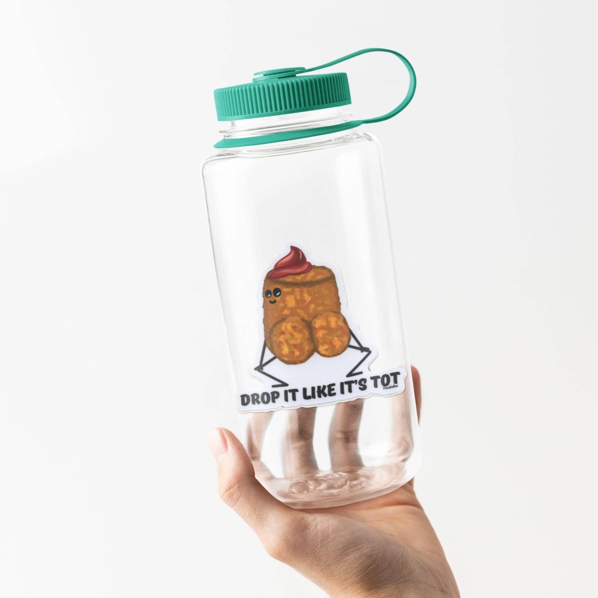 A hand holding a clear plastic water bottle with a green cap and loop handle. The bottle features a cartoon graphic of a smiling tater tot with a red swirl on top and the text 'DROP IT LIKE IT'S TOT' printed below.