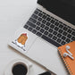 A die-cut sticker featuring a cute cartoon tater tot character wearing a red ketchup swirl on top with the text 'DROP IT LIKE IT'S HOT', shown applied to the palm rest of a silver laptop. The laptop is styled in a flat-lay arrangement on a white wooden desk alongside a cup of black coffee, a white notebook, white earbuds, and an orange spiral notebook.