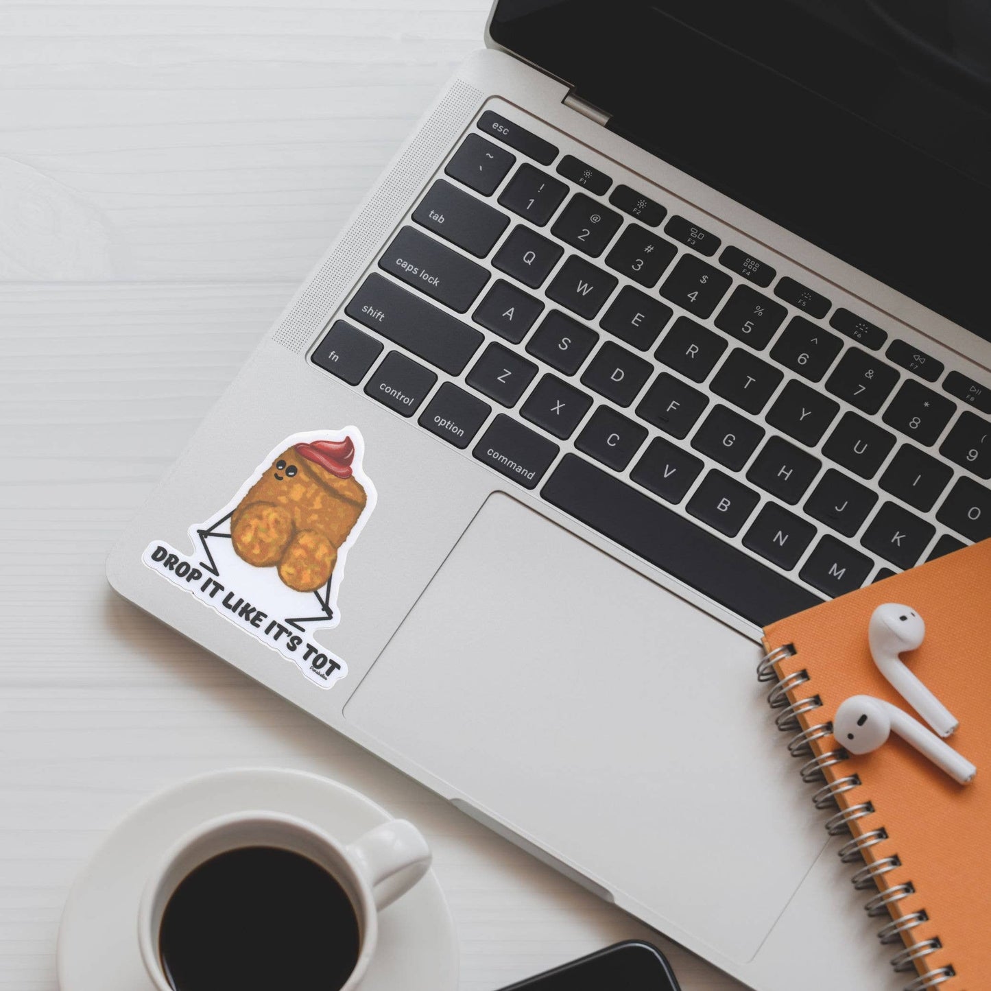 A die-cut sticker featuring a cute cartoon tater tot character wearing a red ketchup swirl on top with the text 'DROP IT LIKE IT'S HOT', shown applied to the palm rest of a silver laptop. The laptop is styled in a flat-lay arrangement on a white wooden desk alongside a cup of black coffee, a white notebook, white earbuds, and an orange spiral notebook.