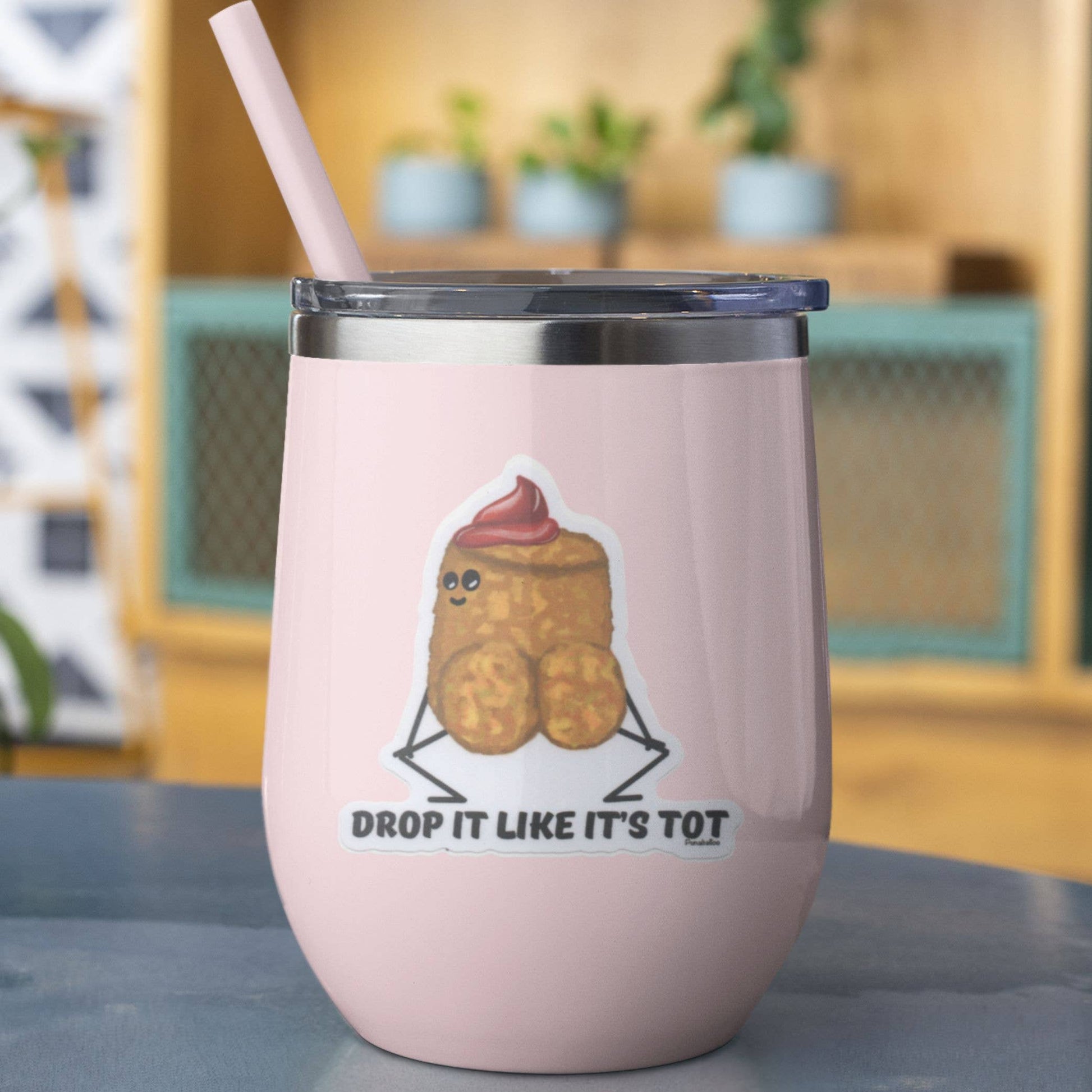A light pink insulated stemless tumbler with a clear lid and straw, featuring a cartoon tater tot graphic and the text 'DROP IT LIKE IT'S TOT' on the front, set against a softly blurred indoor background.