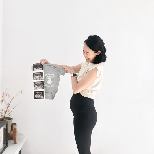 Heather grey baby onesie with white text that reads: Mommy's LIttle Dumpling. Image of a smiling dumpling. Pregnant woman holding ultrasound photos and the baby onesie.