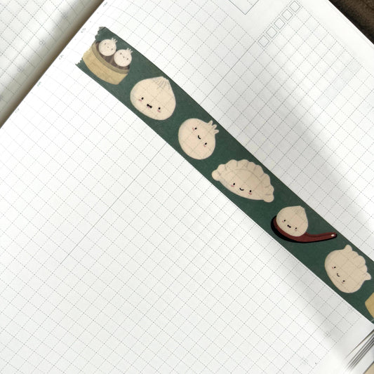 A close-up of a notebook with grid paper featuring a decorative washi tape strip. The tape is dark green with cute cartoon dumpling faces in cream, beige, and white, plus a small red spoon accent.