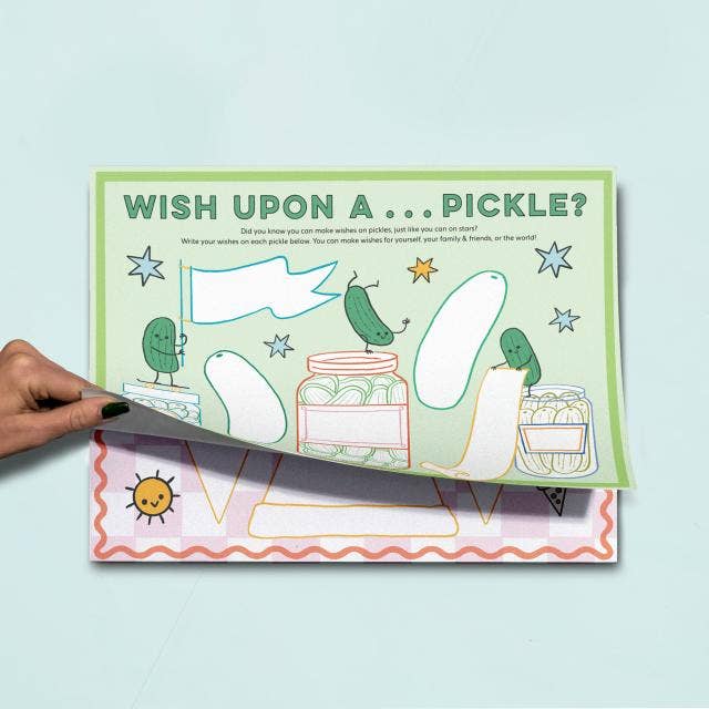 A hand is peeling back a printed activity sheet from another sheet. The top sheet has a light green border and reads 'WISH UPON A ... PICKLE'