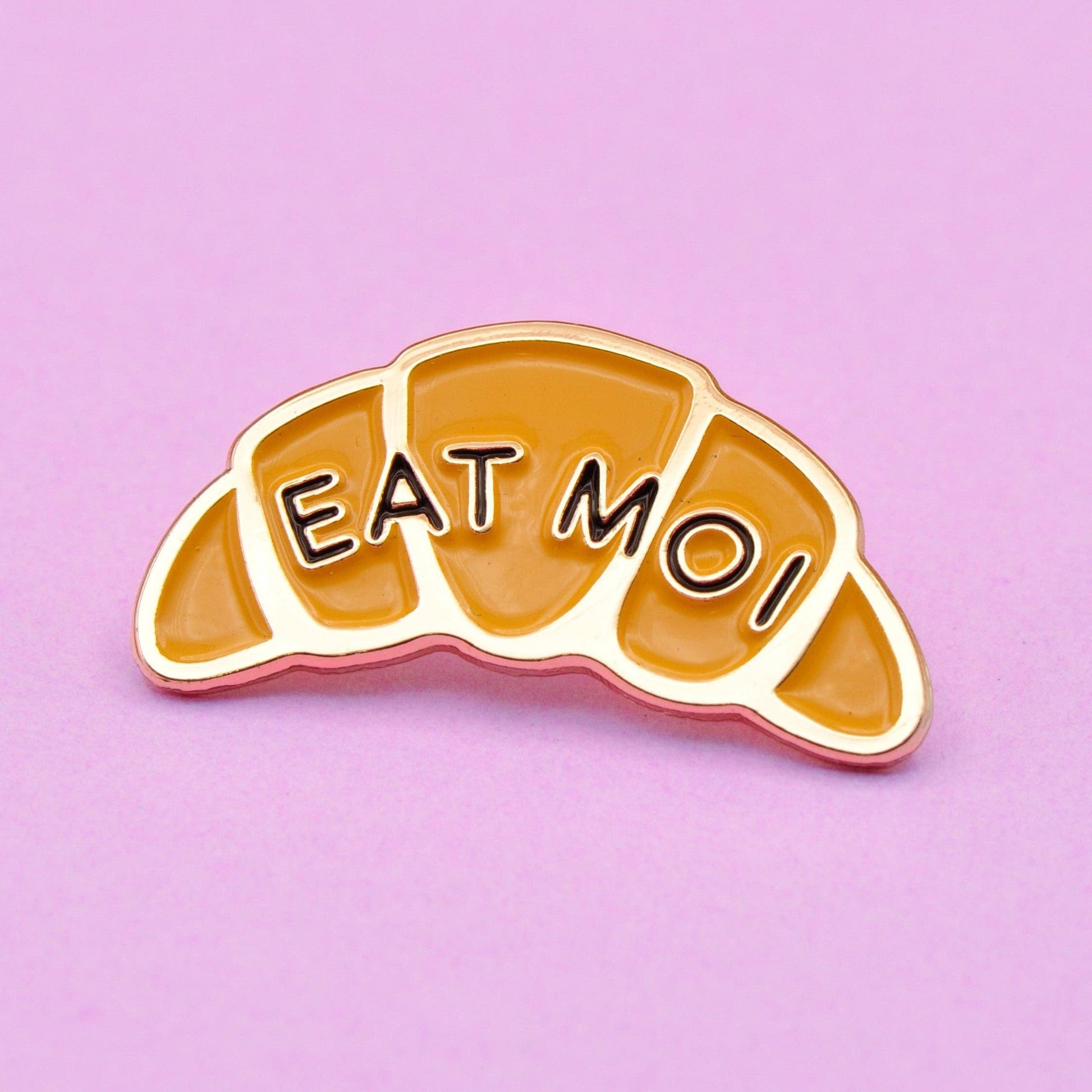 Enamel pin in the shape of a croissant that says "EAT MOI"