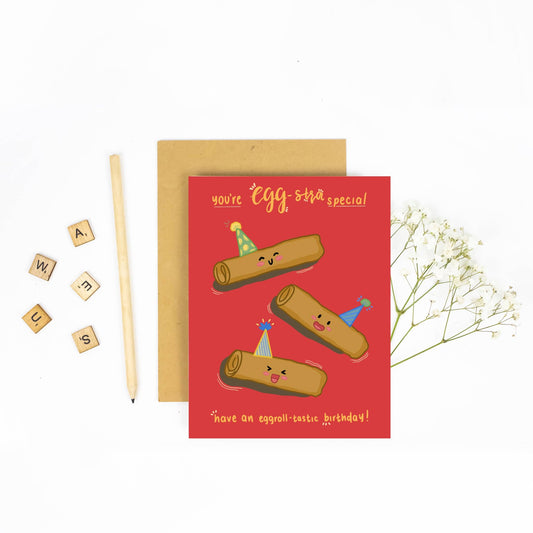 A red greeting card with a brown kraft envelope, styled on a white background with a pencil, Scrabble tiles, and baby's breath flowers. The card reads 'you're egg-stra special' and 'have an eggroll-tastic birthday!' with illustrations of three rolling dough characters wearing party hats.
