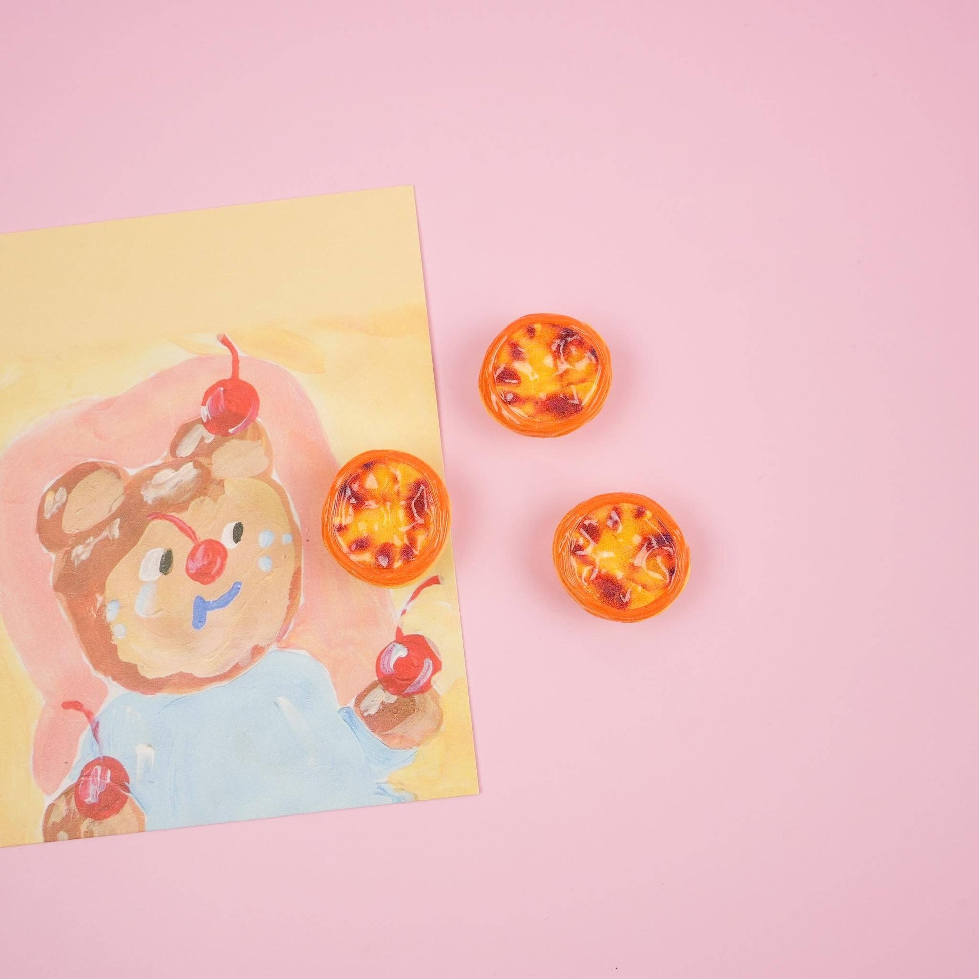 Three egg tart magnets staged on a pink table with a photo of a bear.