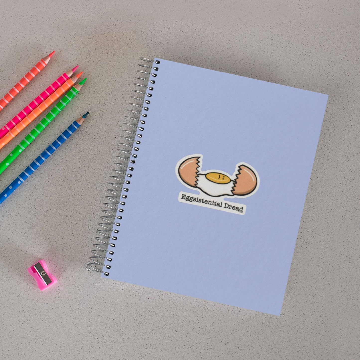 A light blue spiral notebook with a cartoon sticker of a cracked egg and the text 'Eggistential Dread' on the cover, shown on a light gray surface with colored pencils and a pink sharpener nearby.
