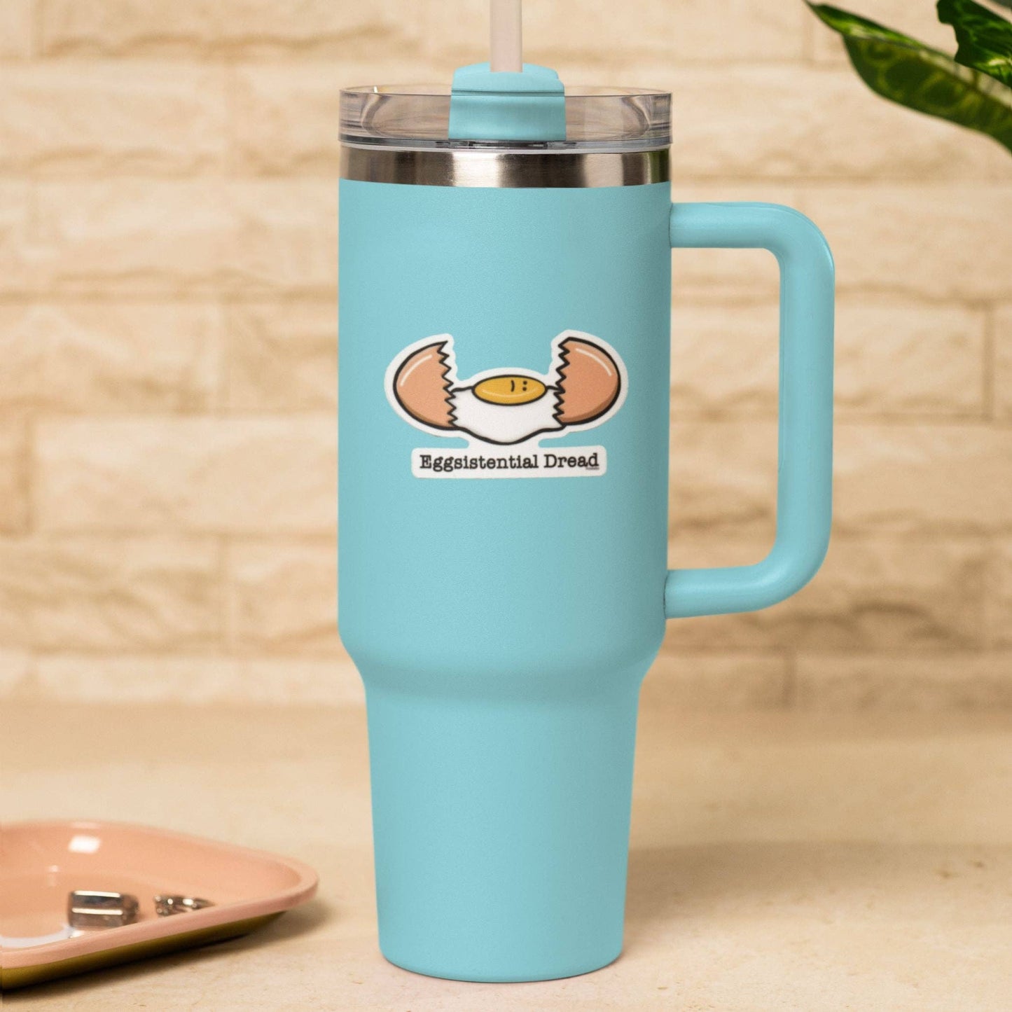 A light blue insulated travel tumbler with a handle, stainless steel rim, clear lid, and straw, featuring a sticker of a cracked egg with the text 'Eggistential Dread' on the front, set against a beige brick wall background.