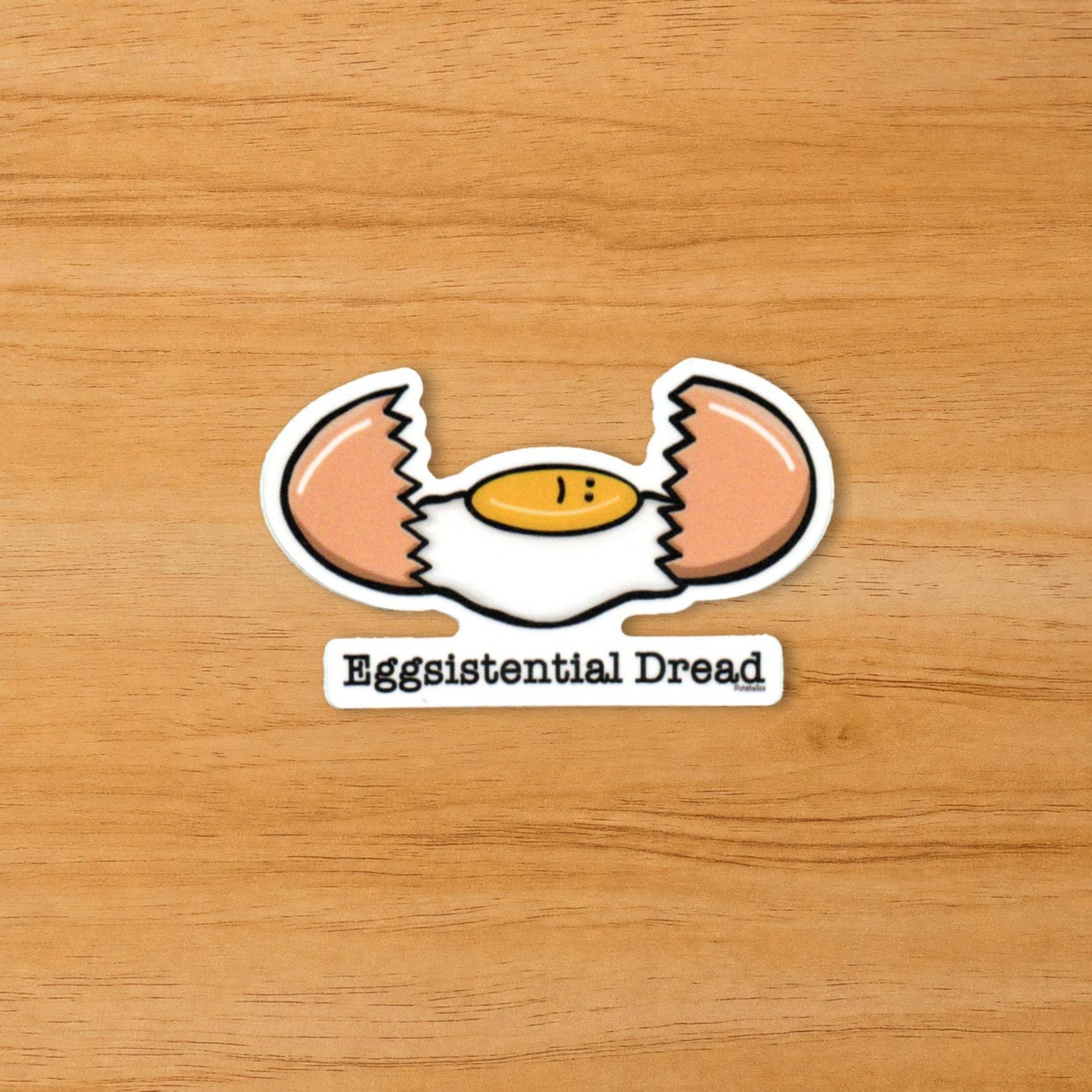 A die-cut sticker featuring a cracked egg with a sad face and the text 'Eggsistential Dread' below it, placed on a light brown wood background.