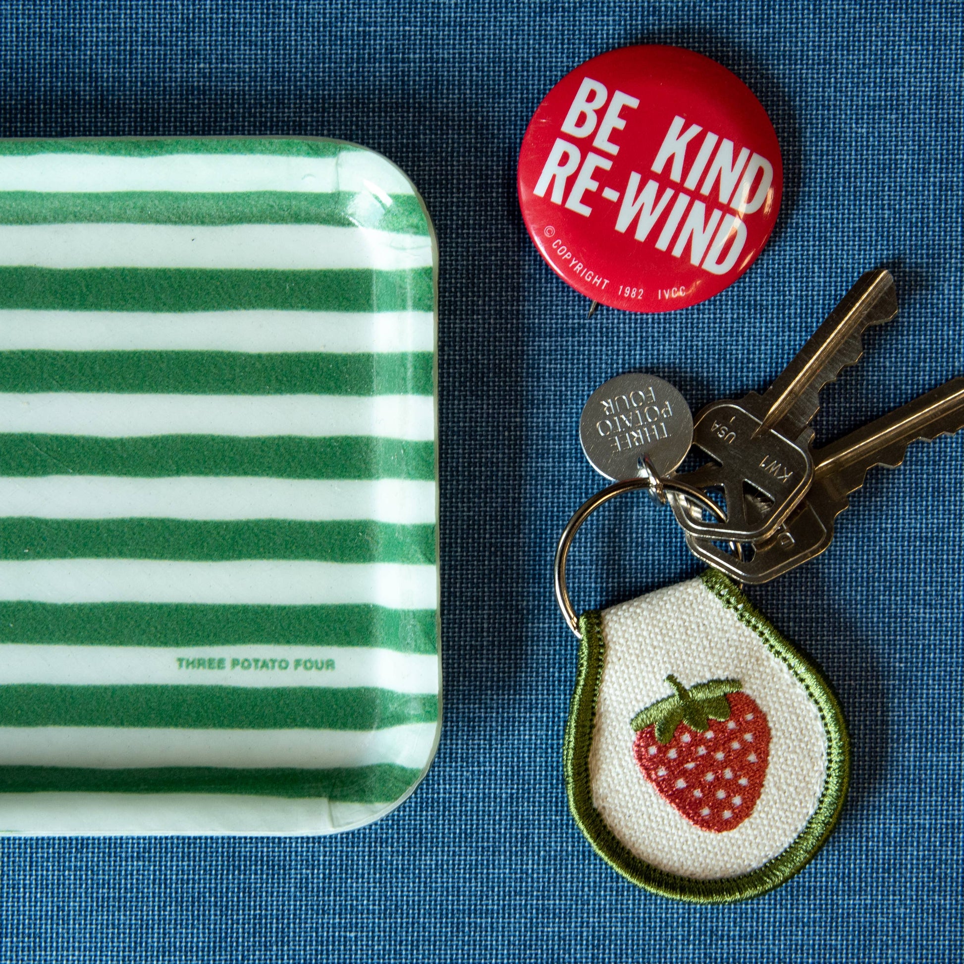 Embroidered Strawberry key chain with a single strawberry and bordered with an army green threading. Metal ring has an extra metal tag that says: Three Potato Four.