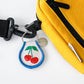 Embroidered Cherry Keychain. Duo cherries with leaves with a blue border. Keyring has an additional metal piece that says: Three Potato Four. Diplayed on a yellow side pack.