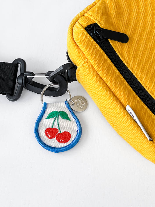 Embroidered Cherry Keychain. Duo cherries with leaves with a blue border. Keyring has an additional metal piece that says: Three Potato Four. Diplayed on a yellow side pack.