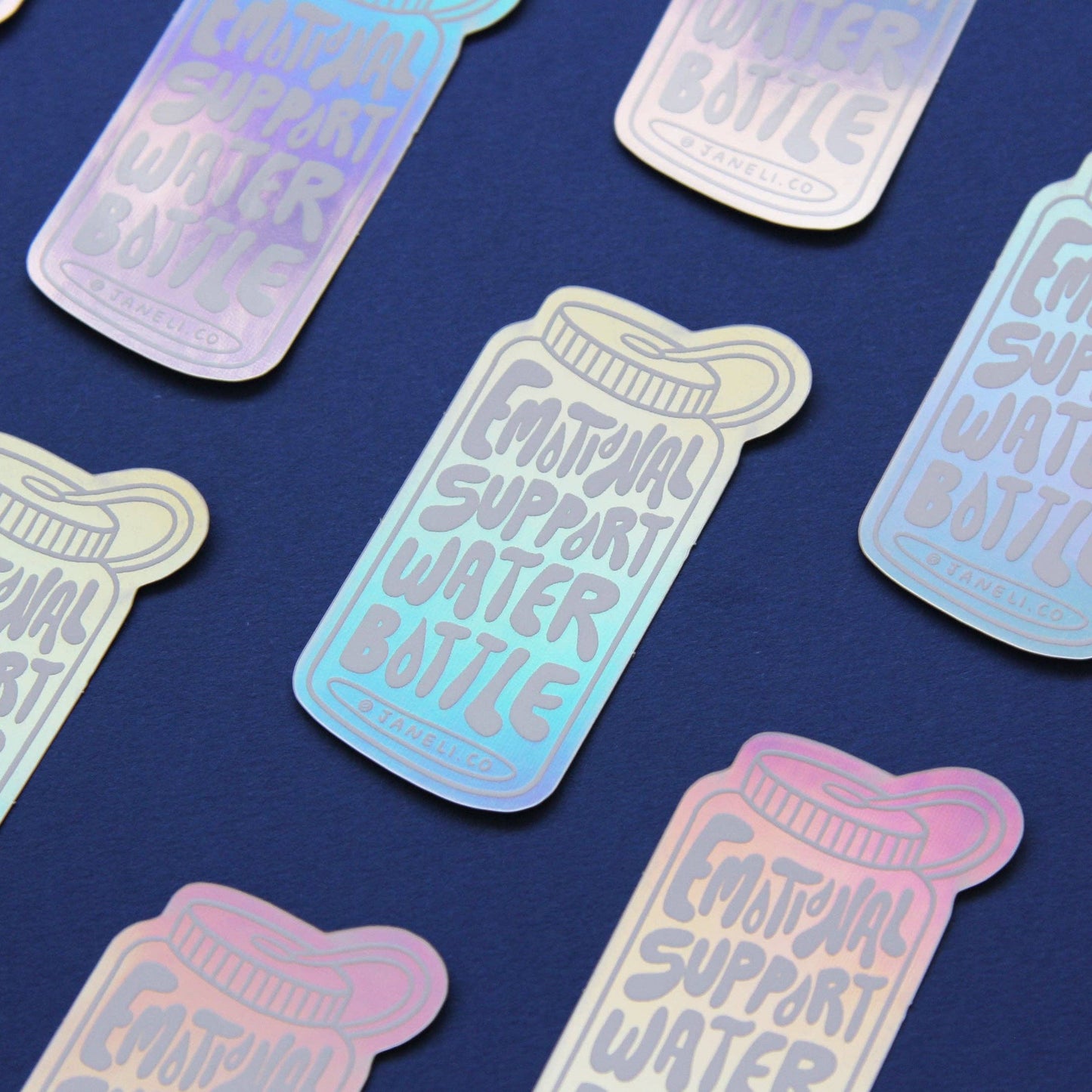 A repeating pattern of die-cut stickers shaped like a water bottle, featuring the text 'EMOTIONAL SUPPORT WATER BOTTLE' in a light gray font over an iridescent pastel gradient background. The stickers are arranged diagonally on a dark navy blue surface.
