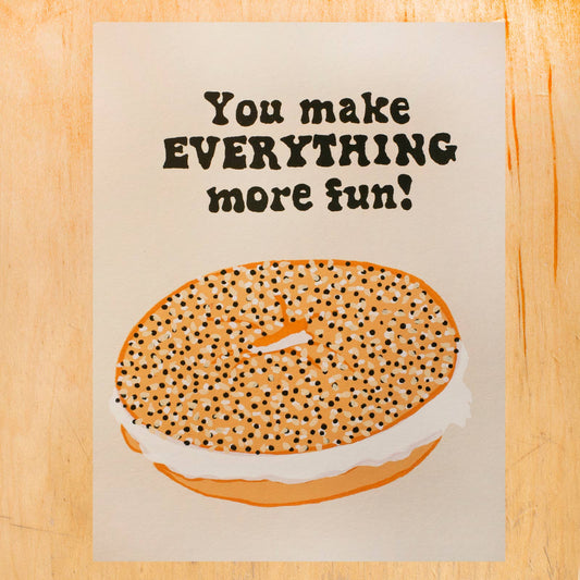 Card with a bagel illustration and text on a wooden surface. Text reads "You make everything more fun!"