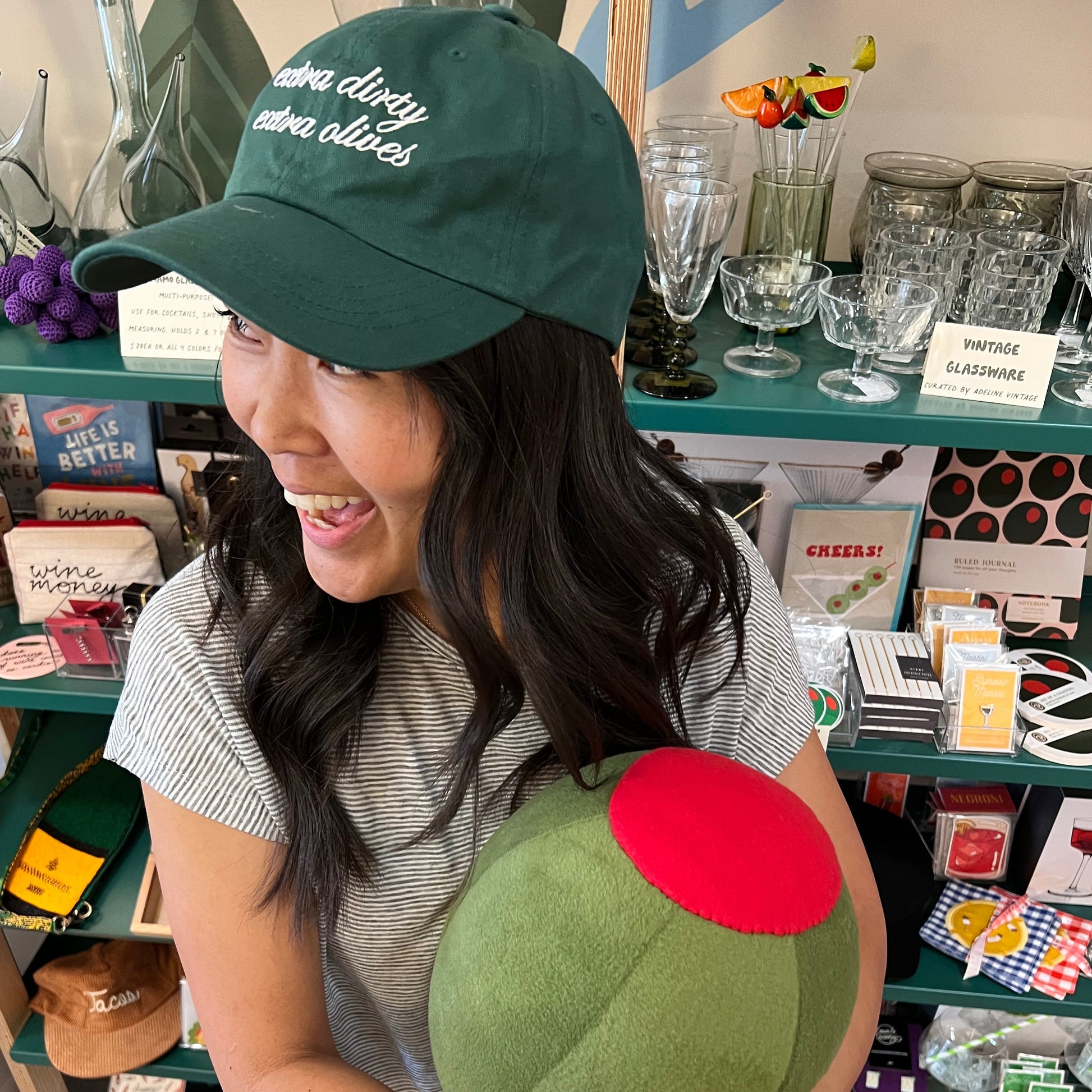green cap with extra dirty extra olives hat worn on a person holding an olive plush pillow