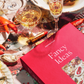 Puzzle called "Fancy Ideas" in a red box. The image of the puzzle is a fancy feast of seafood, caviar, martinis and champagne. Table is staged to show the box, completed puzzle and drinks with lipstick