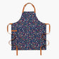 Flat image of dark blue apron with a design pattern of a variety of colorful produce with brown neck and waist straps