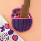 Purple and pink fig-shaped oven mitt held by a hand on a peach background with a white towel featuring fruit designs.