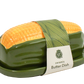 A ceramic butter dish shaped like a corn cob, featuring a glossy dark green base and a bright yellow-orange corn tip, wrapped in a paper band with the brand name 'W' and 'ceramic Butter Dish' printed on it.