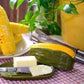 Butter dish with a knife, butter, and corn on the cob on a tablecloth with a plant in the background.