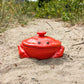 A bright red ceramic crab-shaped lidded container sitting in sand outdoors, with blurred greenery in the background.
