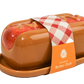 A ceramic butter dish shaped like a hot dog in a bun, with glossy brown glaze, red sauce details, and a red-and-white checkered paper band around the center. An orange label on the band reads 'ceramic Butter Dish' with a monogram logo.