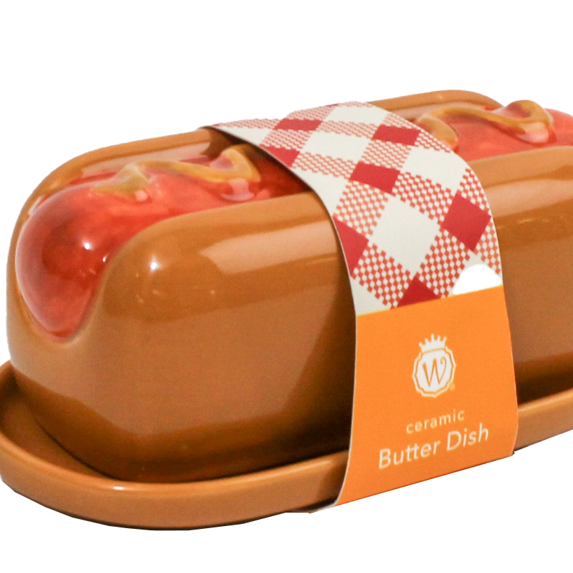 A ceramic butter dish shaped like a hot dog in a bun, with glossy brown glaze, red sauce details, and a red-and-white checkered paper band around the center. An orange label on the band reads 'ceramic Butter Dish' with a monogram logo.