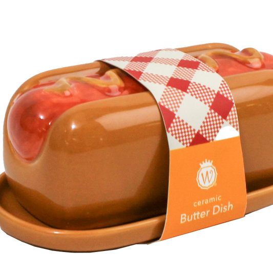A ceramic butter dish shaped like a hot dog in a bun, with glossy brown glaze, red sauce details, and a red-and-white checkered paper band around the center. An orange label on the band reads 'ceramic Butter Dish' with a monogram logo.