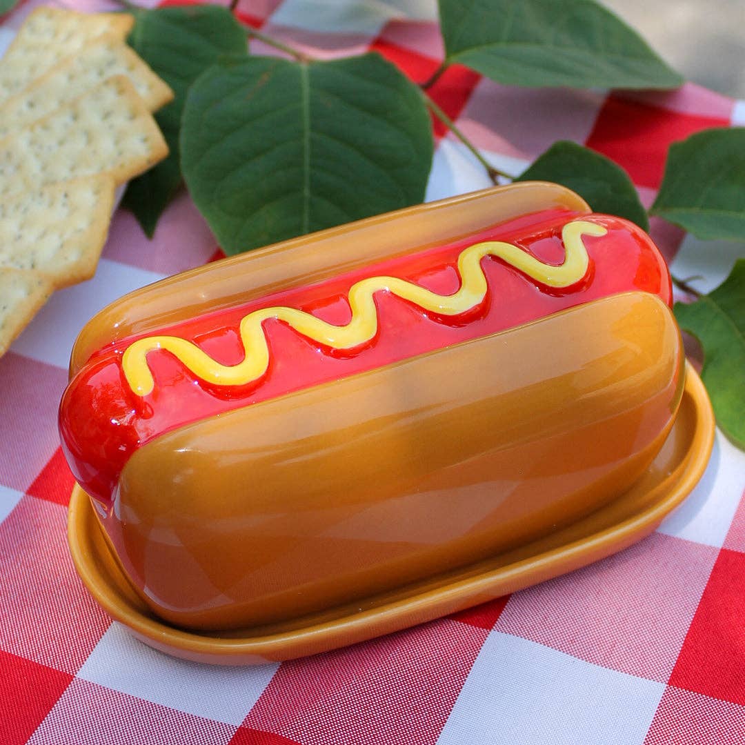 A novelty hot dog-shaped butter dish or container with a glossy orange-brown bun, red filling, yellow mustard line, and matching orange-brown base, styled on a red-and-white checkered picnic blanket with crackers and green leaves.