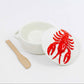 A white ceramic brie baking dish with a matching lid featuring a raised red lobster design, shown next to a small wooden spoon on a clean white background.