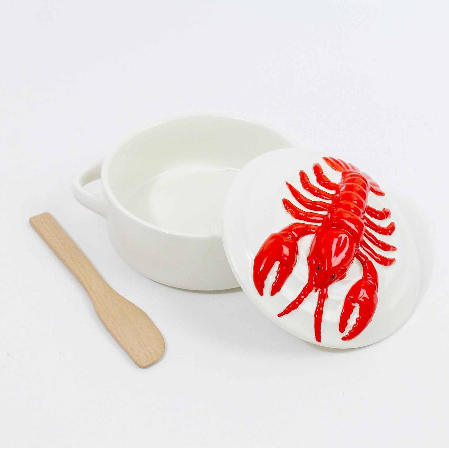 A white ceramic brie baking dish with a matching lid featuring a raised red lobster design, shown next to a small wooden spoon on a clean white background.