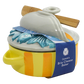 A ceramic brie cheese baker with a yellow and orange striped body, a white lid featuring a blue fish design, and a wooden spoon resting on top, wrapped in a blue paper band.