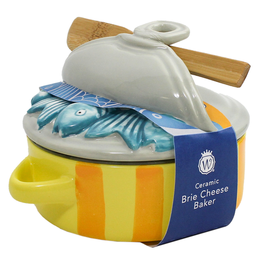 A ceramic brie cheese baker with a yellow and orange striped body, a white lid featuring a blue fish design, and a wooden spoon resting on top, wrapped in a blue paper band.