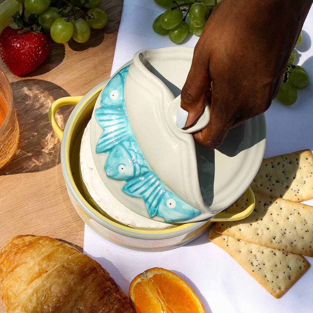 Person opening a ceramic cheese dish with fish design on a table with snacks