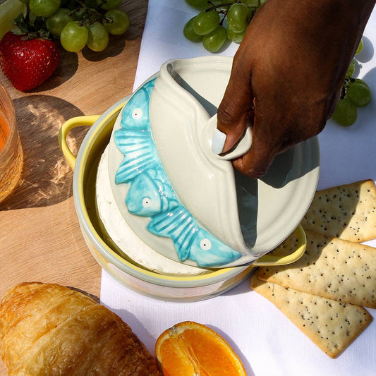 Person opening a ceramic cheese dish with fish design on a table with snacks