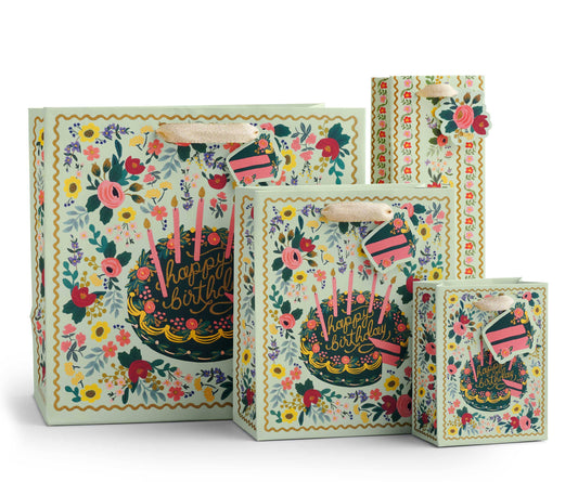 Gift bag shown in assorted sizes. Image on white background - intricate, vintage-inspired floral print surrounding birthday cake decorated with pink and purple flowers, dark frosting base, and pink candles. Squiggly golden yellow border. Very maximalist.