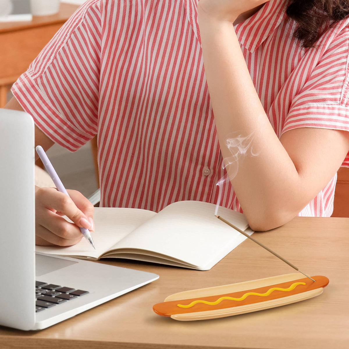 A person in a pink and white striped shirt sits at a wooden desk using a laptop and writing in a notebook, with a hot dog-shaped incense holder on the table holding a smoking incense stick.